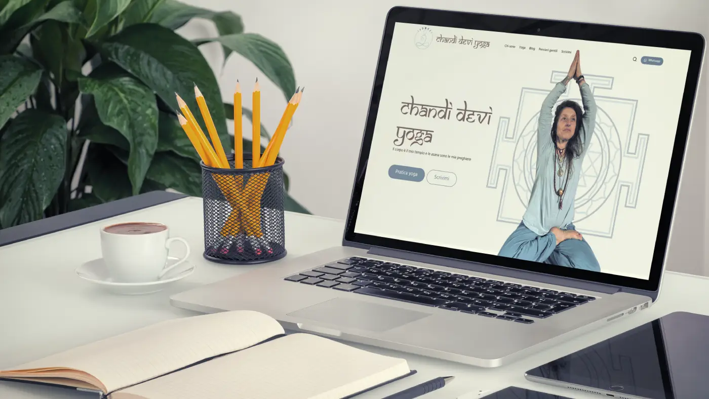 Laptop aperto su scrivania che mostra il sito web di un’insegnante di yoga, accanto a una tazza di caffè, un portapenne con matite gialle e un quaderno aperto.
