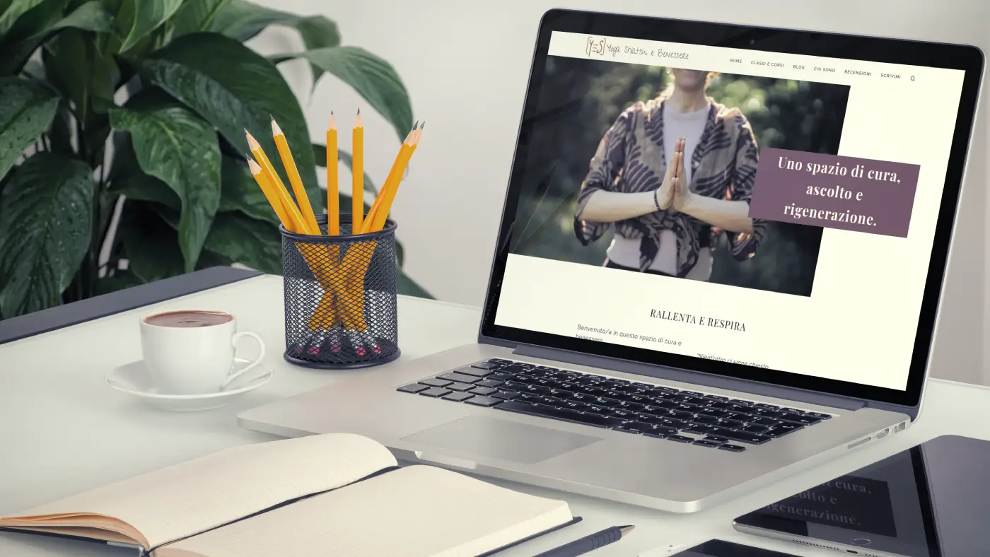 Laptop aperto su scrivania che mostra il sito web di un’insegnante di yoga, accanto a una tazza di caffè, un portapenne con matite gialle e un quaderno aperto.