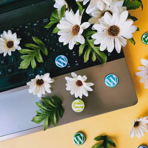Una vista dall'alto su un laptop aperto, parzialmente coperto da fiori artificiali bianchi simili a margherite con centri marroni e foglie verdi. Sulla tastiera e sulla superficie del laptop sono sparse diverse spillette rotonde con il logo di WordPress, sia verdi che blu. Lo sfondo è un tavolo giallo.