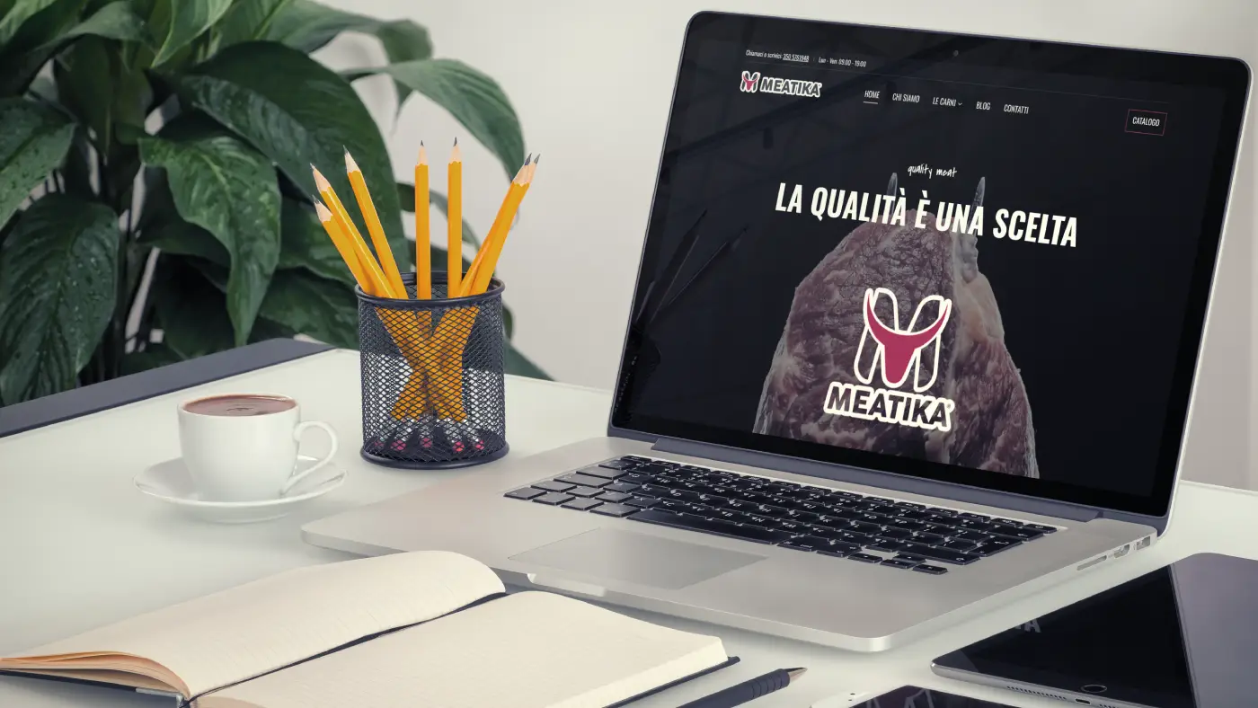 Progetto web per Meatika: il sito è visibile su un laptop in un ambiente da lavoro ordinato, con matite, quaderno e una tazza di caffè a lato.